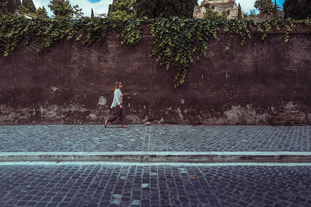 A woman walks along a sunlit cobblestone street in a charming Roman neighborhood, surrounded by pastel buildings with shuttered windows and soft afternoon shadows.