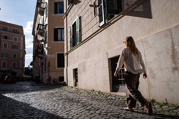 A woman walks along a sunlit cobblestone street in a charming Roman neighborhood, surrounded by pastel buildings with shuttered windows and soft afternoon shadows.