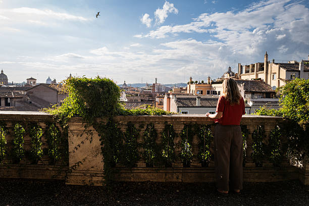A woman along a shaded path at the Pincian Hill (Pincio Terrace) in Rome, with a panoramic view of the city's historic domes and rooftops in the background on a sunny day.
