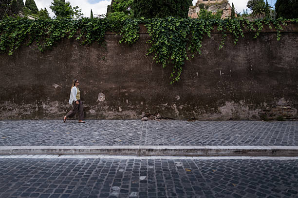 A woman walks along a sunlit cobblestone street in a charming Roman neighborhood, surrounded by pastel buildings with shuttered windows and soft afternoon shadows.