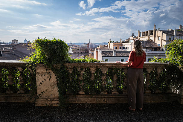 A woman along a shaded path at the Pincian Hill (Pincio Terrace) in Rome, with a panoramic view of the city's historic domes and rooftops in the background on a sunny day.