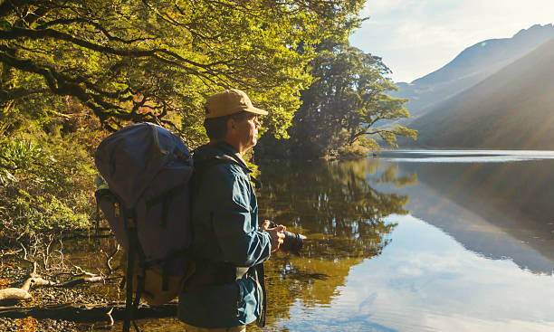 Male hiker wearing a backpack and a hat holding camera looking at breathtaking view of mirror lake with mountain reflection in water.