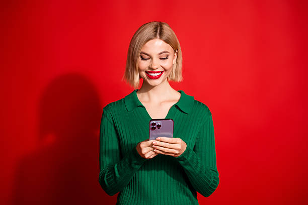 istock Charming young woman smiling while using smartphone against vibrant red background wearing a stylish green sweater 2216397504