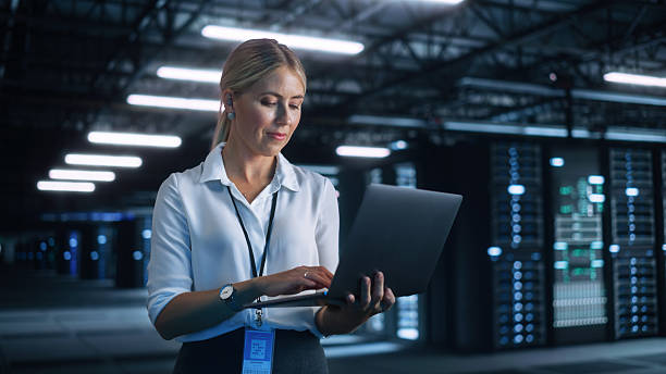 istock Female Data Center IT Technician Standing at the Server Rack Corridor with Laptop Computer. She is Visually Inspecting Something while Looking at the Screen. Night Office Concept 2216425259