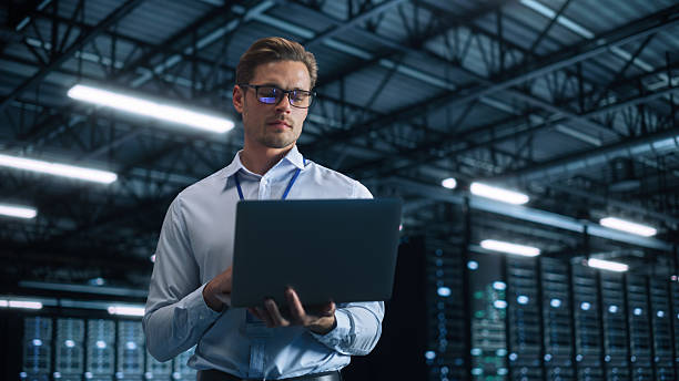 istock Male IT Engineer Working with Laptop in Data Center. Technician or Developer Standing at the Server Rack Corridor with a Laptop Computer. High-Speed Data Transfer, Analytics, Statistics Concept 2216425407