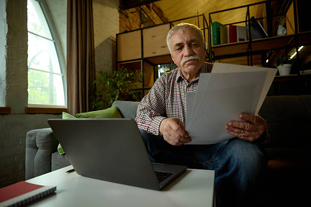 istock Focused elderly man with mustache sitting on sofa in modern living room, looking at papers, with laptop on table 2216426120