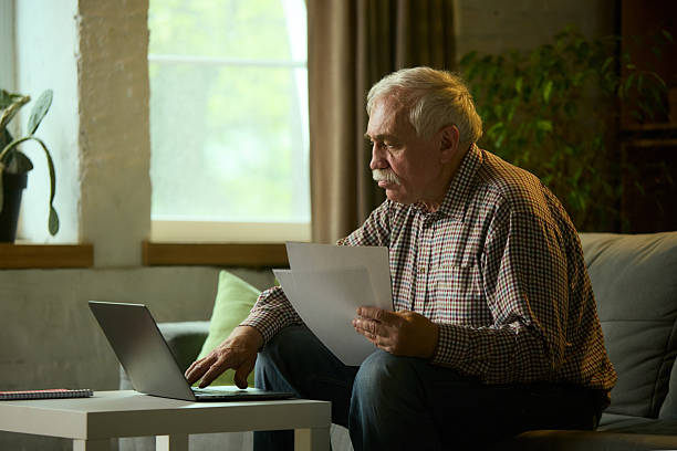 istock Focused elderly man with mustache sitting on sofa in modern living room, looking intently at laptop, holding papers and studying. 2216426123