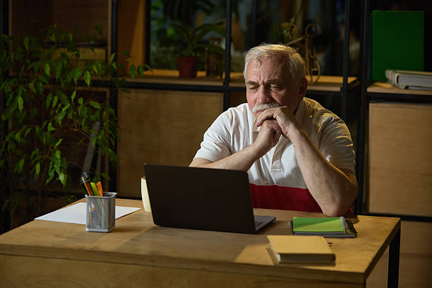 istock Focused elderly man sitting at wooden desk, using laptop and notebooks in quiet home office. Digital learning, modern technologies 2216426175