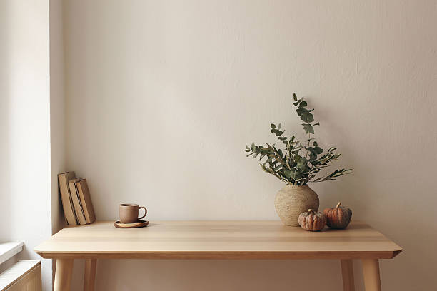 istock Autumn interior still life. Vase with eucalyptus, olive tree branches, pumpkins. Cup of tea, coffee, books. Wooden table at window. Home office. Minimal Scandi interior. Empty white wall background. 2216428765