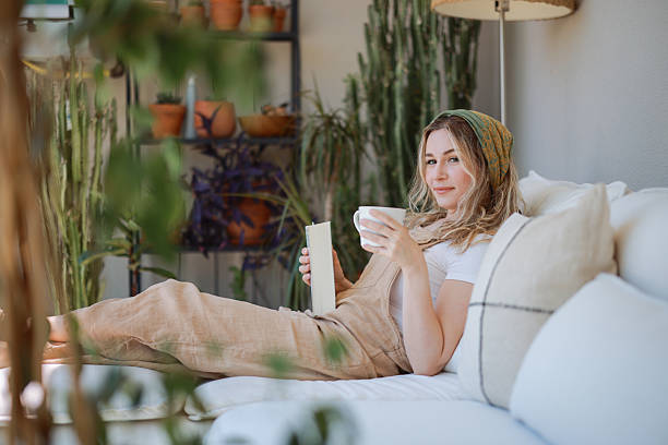 istock Young woman in overalls sitting on a sofa reading a book and drinking coffee 2216473768