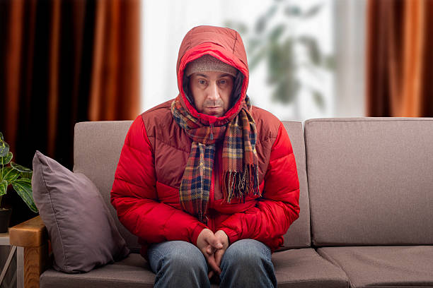 istock Middle age male model in red parka and checkered scarf getting cold sitting on the sofa in cold living room 2216487399