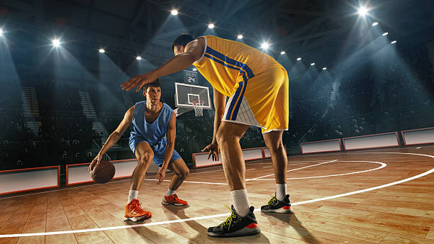 istock Composite collage. Basketball player in blue dribbles low, facing defender in yellow uniform on 3D basketball court with visible hoop and audience behind. 2216491599