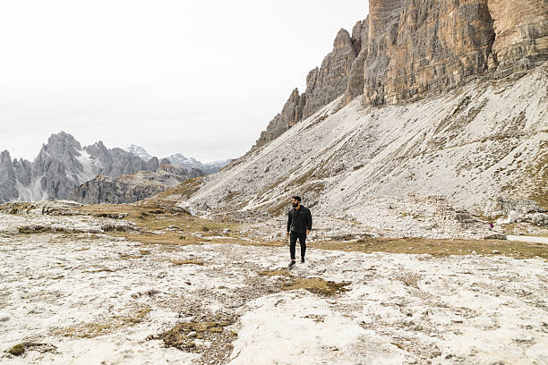 Man hiking through the dramatic rocky landscape of the Dolomites, northern Italy. The traveler is wearing a black puffer jacket and black pants, walking along a rugged mountain trail surrounded by steep limestone cliffs and alpine terrain. A scenic backdrop of mountains and valleys enhances the raw beauty of this UNESCO World Heritage site.