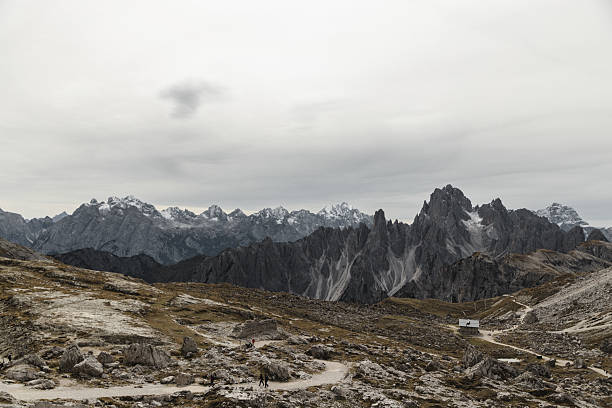 A small mountain hut surrounded by the dramatic rocky formations of the Dolomites in Italy. The landscape features a clear hiking trail leading through the rugged terrain, with snow-covered peaks visible in the distance.