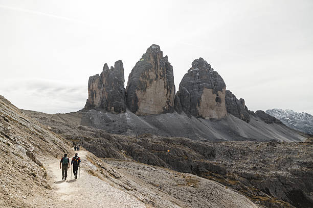 Two hikers walking along a mountain trail towards the famous Tre Cime di Lavaredo peaks in the Dolomites, Italy. The rugged landscape and dramatic rock formations create an impressive backdrop for outdoor adventures.