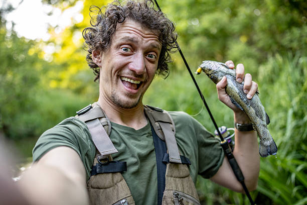 A joyful fisherman takes a selfie, proudly holding a fresh trout catch against a scenic green forest background, embodying excitement and success in the tranquil pursuit of fishing activities.