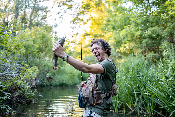 A fisherman excitedly displays a freshly caught fish while standing in a lush, wooded stream landscape, capturing the thrill of outdoor adventure and the joy of fishing in nature.