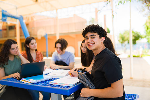 Portrait of a smiling male teenager student taking notes and studying with classmates using laptop and books in a high school campus