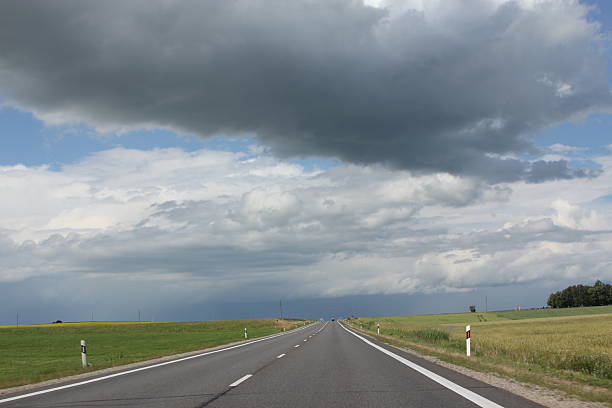 A long empty road disappears into the horizon as storm clouds gather above the green European fields. The contrast between sunlight and ominous clouds creates a compelling atmosphere of tension and solitude.