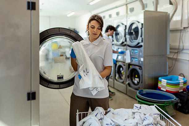 A focused laundry worker in protective gloves handles freshly laundered linens in a commercial laundry room equipped with industrial washing machines. The scene emphasizes cleanliness and efficiency in a professional setting.