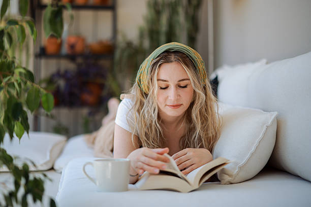 istock Young woman lying on a sofa reading a book and drinking coffee 2216550049