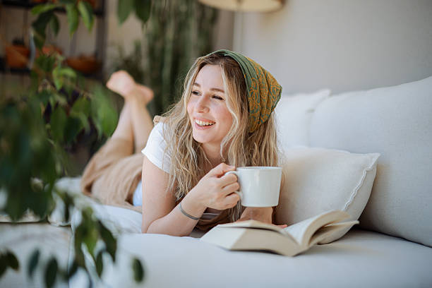 istock Young woman lying on a sofa reading a book and drinking coffee 2216550424
