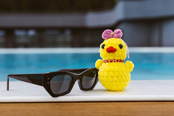 A knitted yellow chick with cool black sunglasses rests by a pool. The scene captures a relaxed, summery vibe vacation.