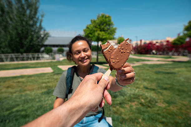 Two individuals share a cheerful moment in a park, holding ice cream bars. The sunlit environment and bright colors highlight friendship, summertime joy, and relaxation outdoors.