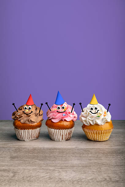 A trio of cupcakes with googly eyes, smiles and party hats on a purple background ready for a celebration!
