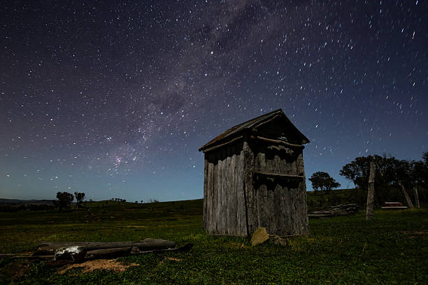 A brilliant arc of the Milky Way stretches through the night sky, surrounded by a dense field of stars. In the foreground, the stark silhouette of an outhouse adds depth