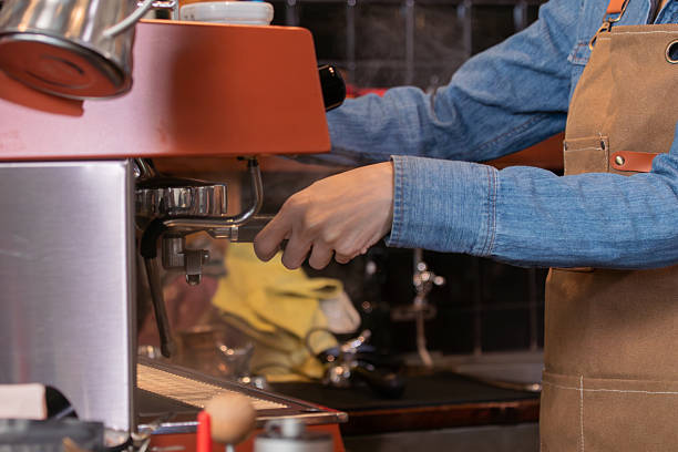 Unrecognizable hands of Asian barista female making hot coffee from professional machine standing behind cafe shop counter bar, young waitress worker wearing apron working in cozy small restaurant