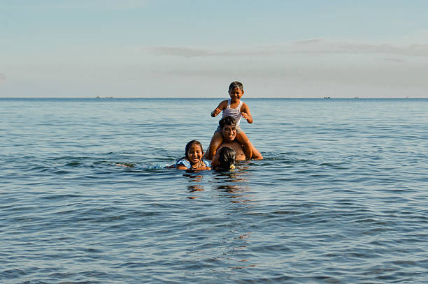 At the beach, a cheerful boy on dadG??s shoulders while mom gently holds their daughter in the sea.