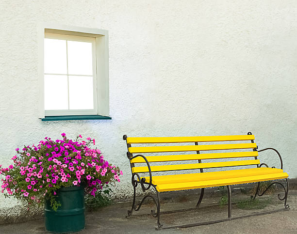 istock Yellow bench under the window on a light wall with copyspace with pink flowers in pots 2216606651