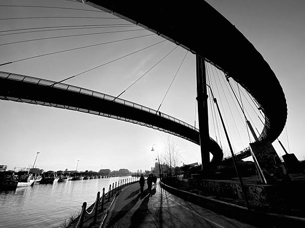 Black and white photo of the Ponte del Mare, a modern pedestrian bridge over the Pescara River, with fishing boats docked in the background