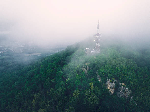 Rows of dense green forests cover the hills, creating a picturesque mountainous landscape under a bright blue sky with white clouds. On the horizon, through the treetops, silhouettes of ancient rocks emerge.