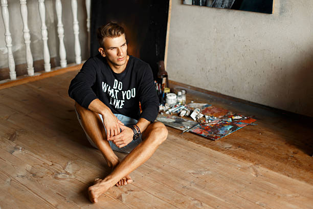 Young handsome guy in a black pullover and denim shorts sitting on the floor in a studio, Artist
