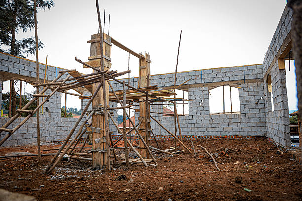 construction phase of a residential house, featuring two central support pillars erected for enhanced structural integrity. Surrounding these pillars are 99 concrete blocks, each laid with precision as part of a strong and enduring foundation. The use of wooden scaffolding and freshly dug earth highlights the craftsmanship and labor behind building a secure home — a future space of safety, warmth, and belonging.