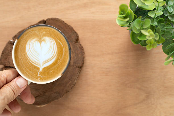 Hand hold hot latte coffee with art heart shape in ceramic cup and little tree on wooden background. Top view, Concept for a banner, cafe, or coffee menu.