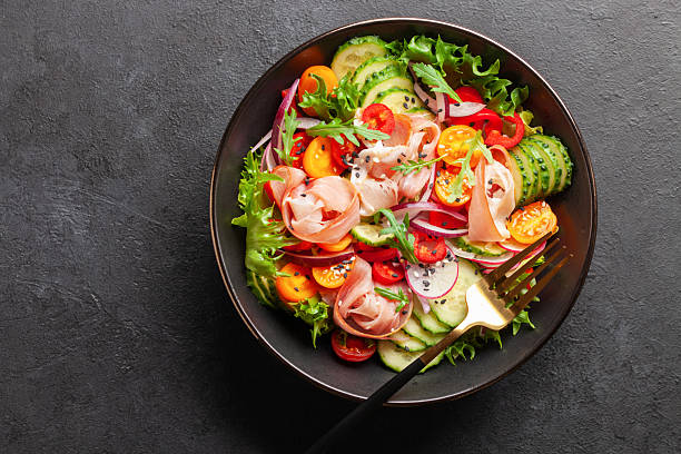 istock Salad with fresh vegetables and jamon in a black bowl on the table. Top view 2216660872
