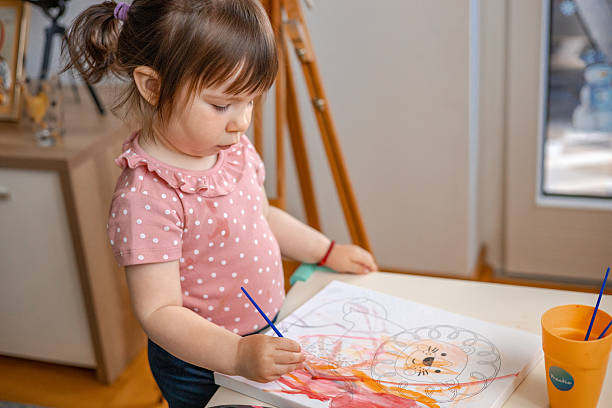 istock Young girl wearing a pink blouse paints a lion outlined on canvas at a small table, using watercolors to bring it to life in her own unique and imaginative style 2216662346