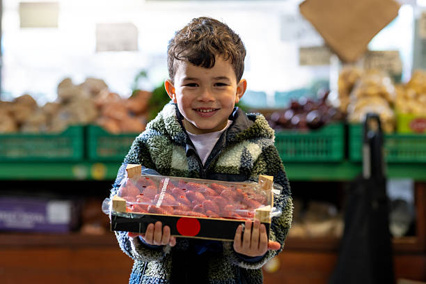 A medium shot of a young boy standing and holding a crate of strawberries from a fruit and vegetable grocer in Whitley Bay. He is looking directly at the camera and smiling. Other fruit and vegetables can be seen in the background.\n\nVideos are available similar to this scenario.
