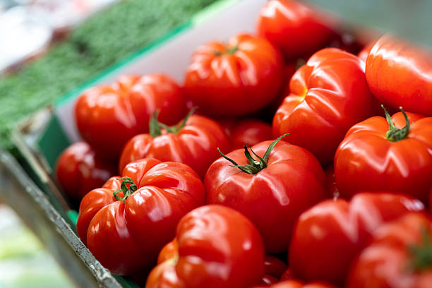 A close-up shot of a crate filled with bright red tomatoes stacked on top of each other. The tomatoes are ripe, vibrant, and ready for sale at a local fruit and vegetable grocer.\n\nVideos are available similar to this scenario.