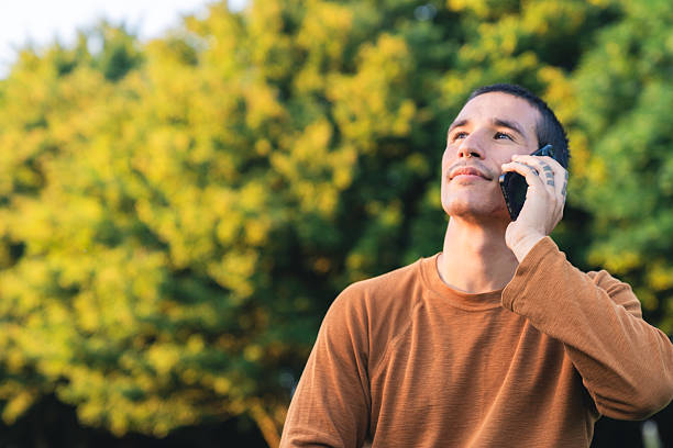 Young man wearing a brown sweater and looking up while making a phone call in a park during golden hour