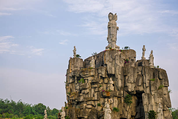 The image presents a striking view of a Guanyin statue, a symbol of mercy and compassion in Buddhism, standing tall against a clear blue sky. The statue, which is the focal point of the image, is depicted with a golden halo and is dressed in flowing white robes, holding a lotus flower, which is often associated with purity and enlightenment. The perspective is from below, looking up towards the statue, which emphasizes its grandeur and the sense of reverence it inspires. The clear sky provides a serene backdrop, enhancing the statue's ethereal beauty and the spiritual ambiance of the scene. This statue, likely located in a place of worship or a sacred site, invites contemplation and serves as a beacon of hope and peace for those who visit.