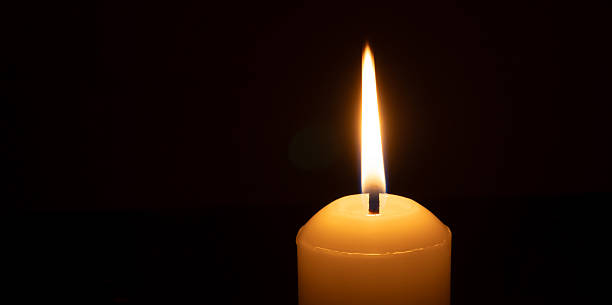 istock A single burning candle flame or light glowing on a big white candle isolated on black or dark background on table in church for Christmas, funeral or memorial service 2216691895