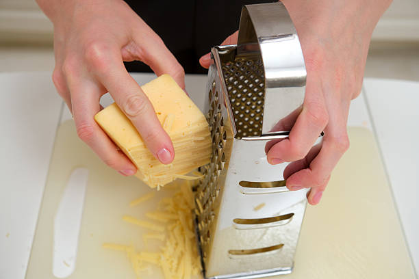 istock Close-up of hands grating cheddar cheese using a stainless steel box grater on a white cutting board in a kitchen. 2216692429