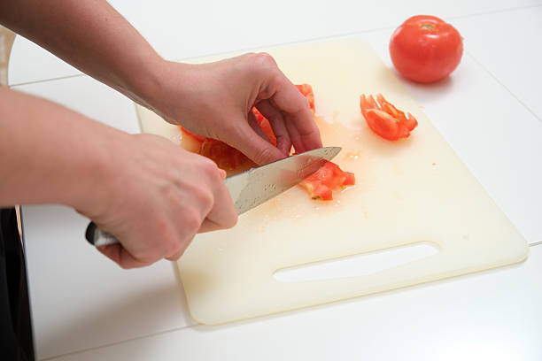 istock Hands preparing food by chopping a juicy red tomato on a plastic board with a sharp knife in a home kitchen setting. 2216692435