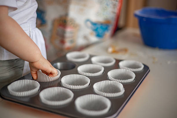 Tiny hands of a young child place paper liners into a baking pan, creating a delightful and detailed moment in the kitchen.