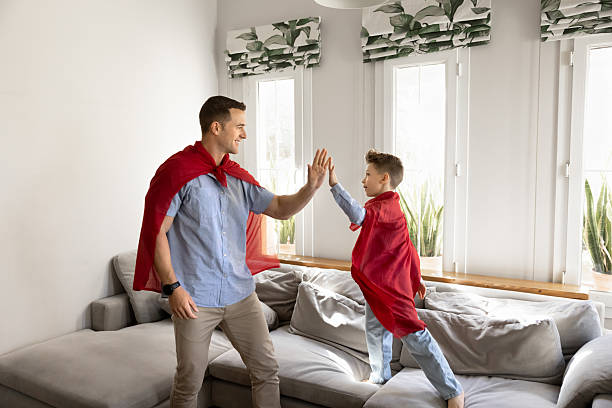 Positive caring dad and playful son kid in red handmade superhero mantles playing active comics game in home living room, giving high five. Father enjoying leisure, family playtime with child