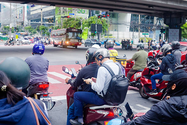 Traffic scene at intersection in Bangkok midtown. Scene is at stoplight of road intersection Rama 9 Road with Thanin Asok / Ratchdaphisek Road on southern side of junction, Traffic is ruling towards north in Bangkok midtown. Many people are using mobile phone while waiting. Some wear crash helmets and some not. Over junction is overpass road of Rama 9 Road. In right background are G Tower and Central Rama 9 mall